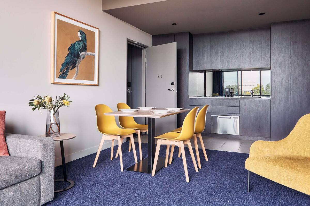 A modern interior with a dining table, yellow chairs, a grey sofa, a blue carpet, and a large window.