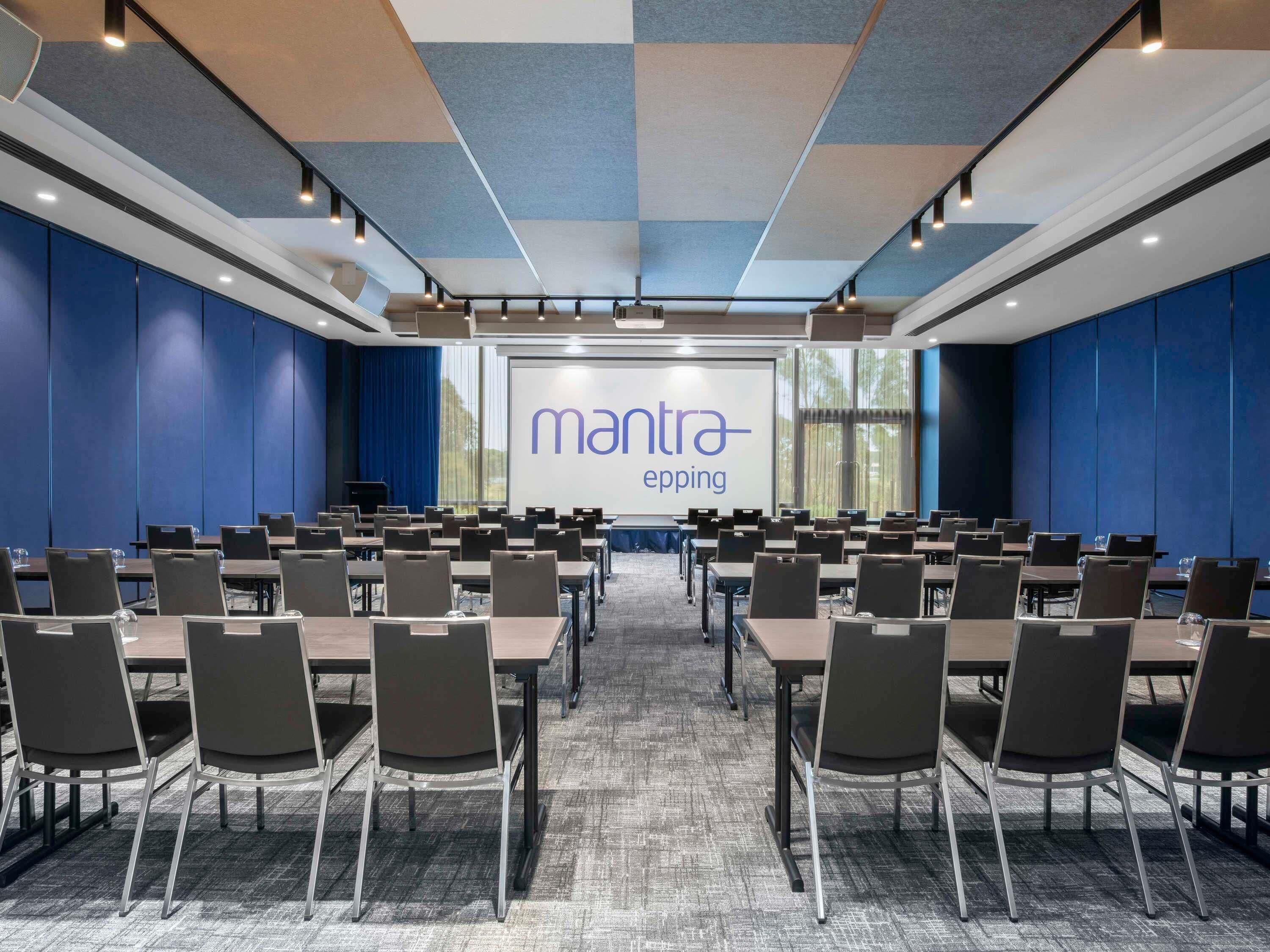A conference room with a large screen displaying 'mantra-eping,' rows of tables and chairs, and a carpeted floor.
