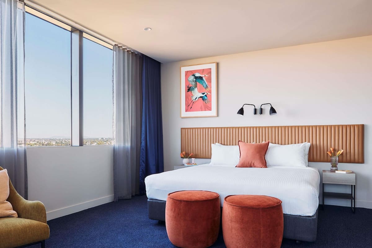 A hotel room with a large bed, two ottomans, a nightstand, a lamp, and a painting.
