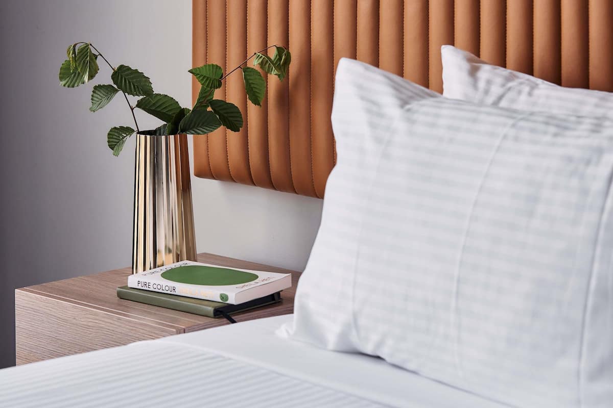 A neatly made bed with a white pillow and a green book titled 'PURE COLOUR' on a wooden nightstand.