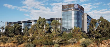 A modern hotel with a curved glass facade, surrounded by lush greenery and a reflective water body.