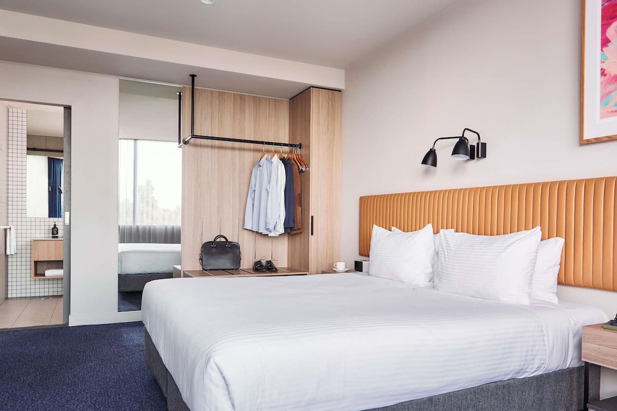 A modern hotel room with a large bed, a wardrobe, a mirror, and a bedside table.