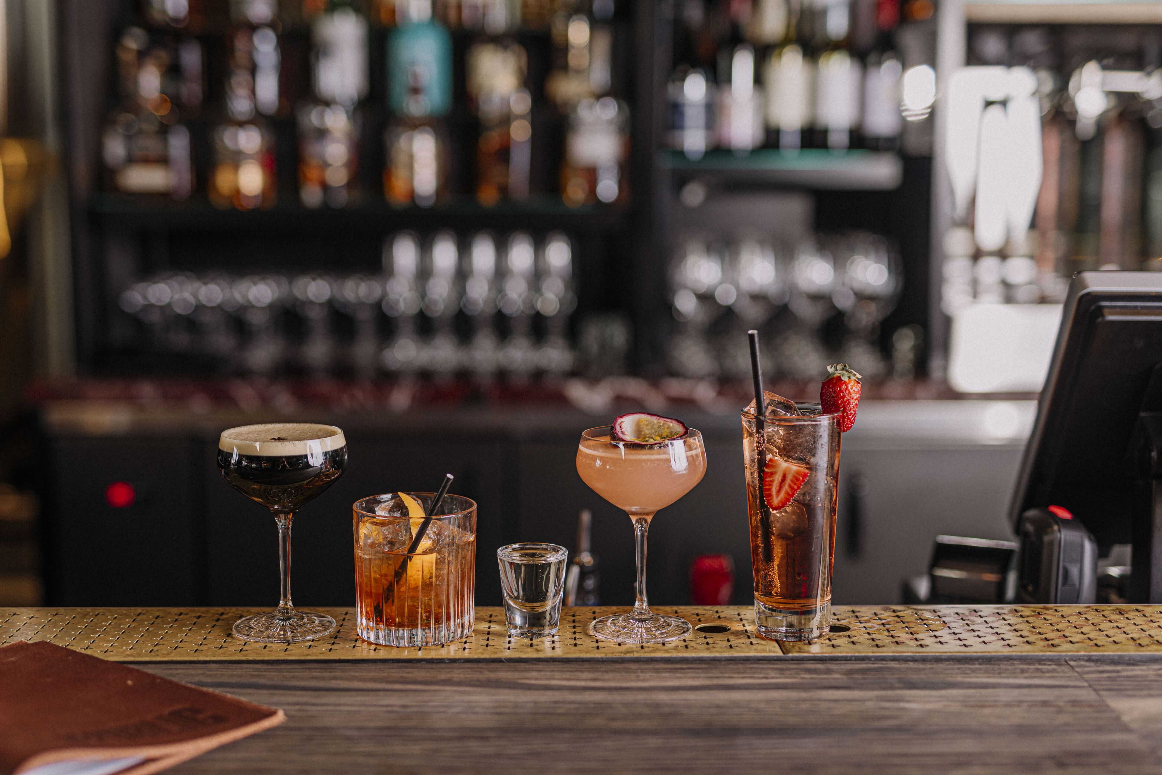 Un bar con diversas bebidas sobre una mesa, un menú y una barra con botellas al fondo.