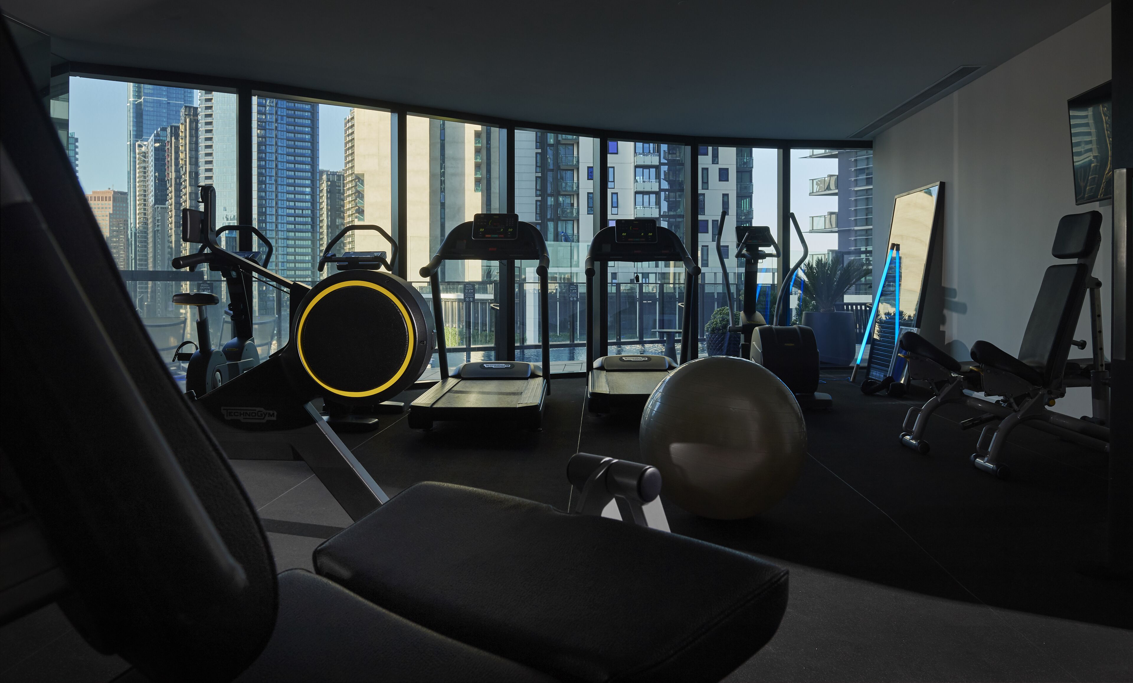 A gym with treadmills, elliptical machines, and exercise balls, featuring large windows with a city view.