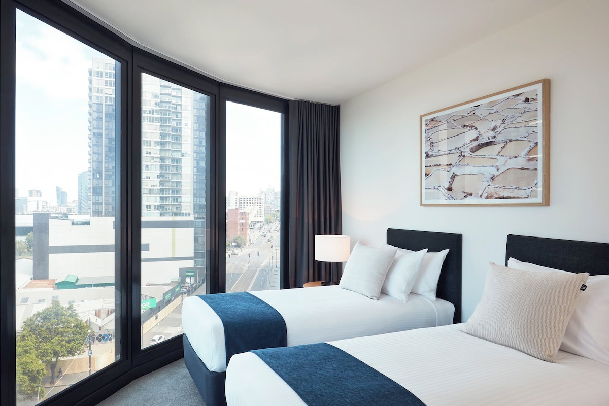 A hotel room with two beds, a large window with a city view, a lamp, and a framed picture on the wall.