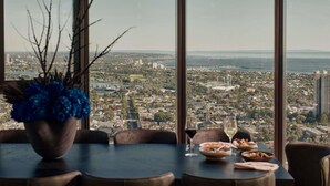 Una tavola da pranzo con vista sulla città attraverso ampie finestre, un vaso con fiori blu e un lampadario.