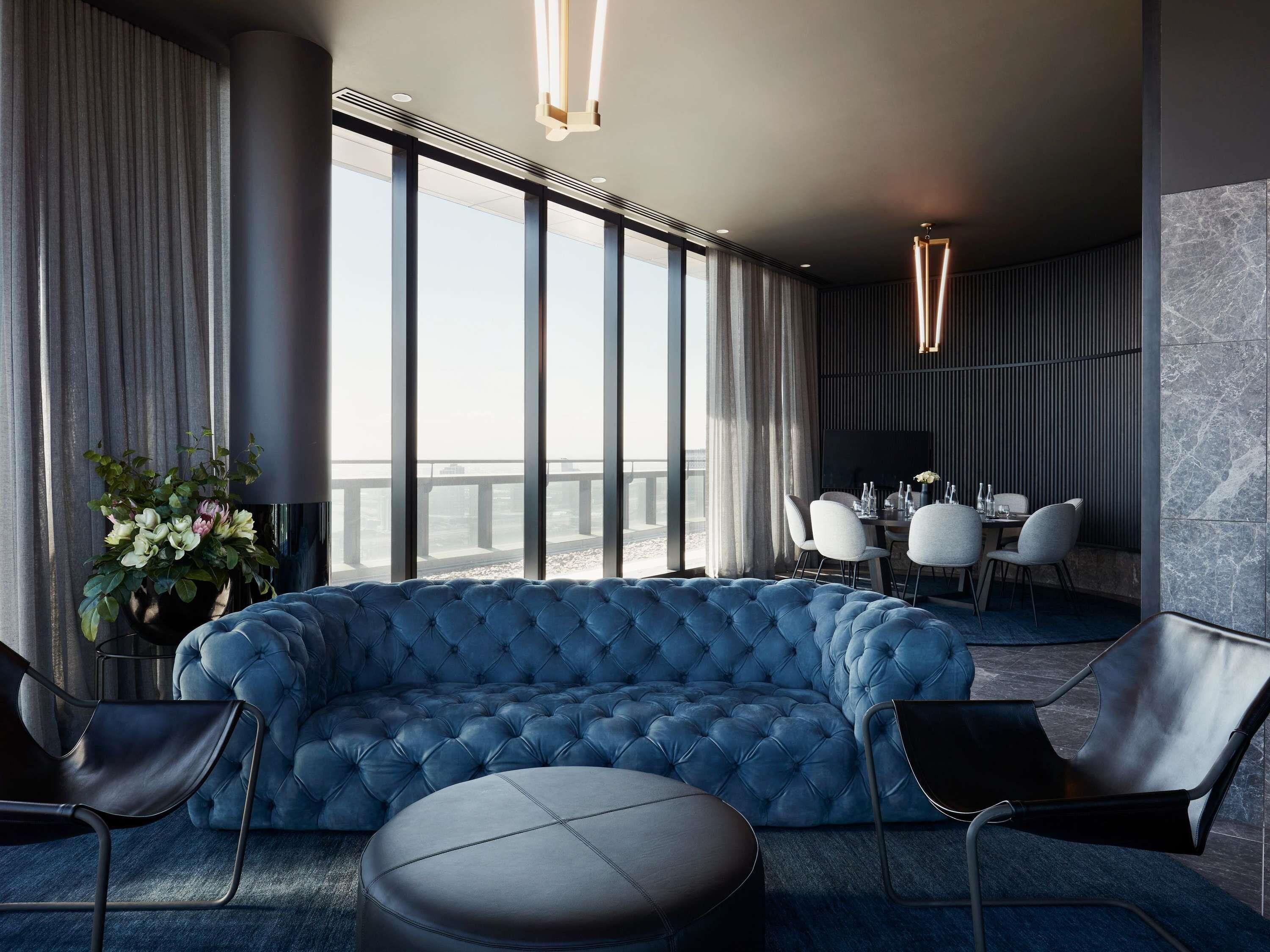 A modern hotel room with a large blue tufted sofa, a dining area with a table and chairs, and large windows offering a view of the city.