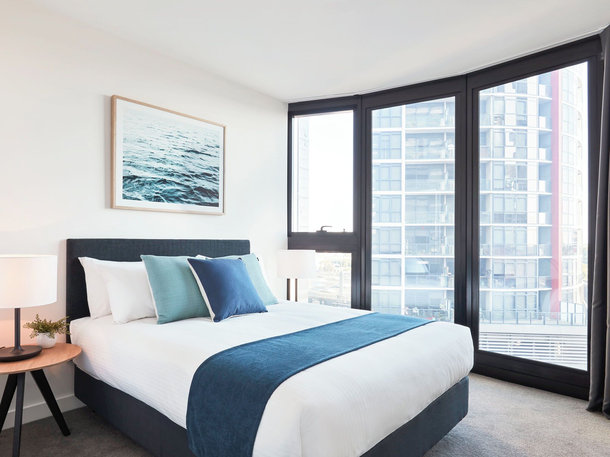 A modern hotel room with a large bed, a view of the city through floor-to-ceiling windows, and a framed picture on the wall.