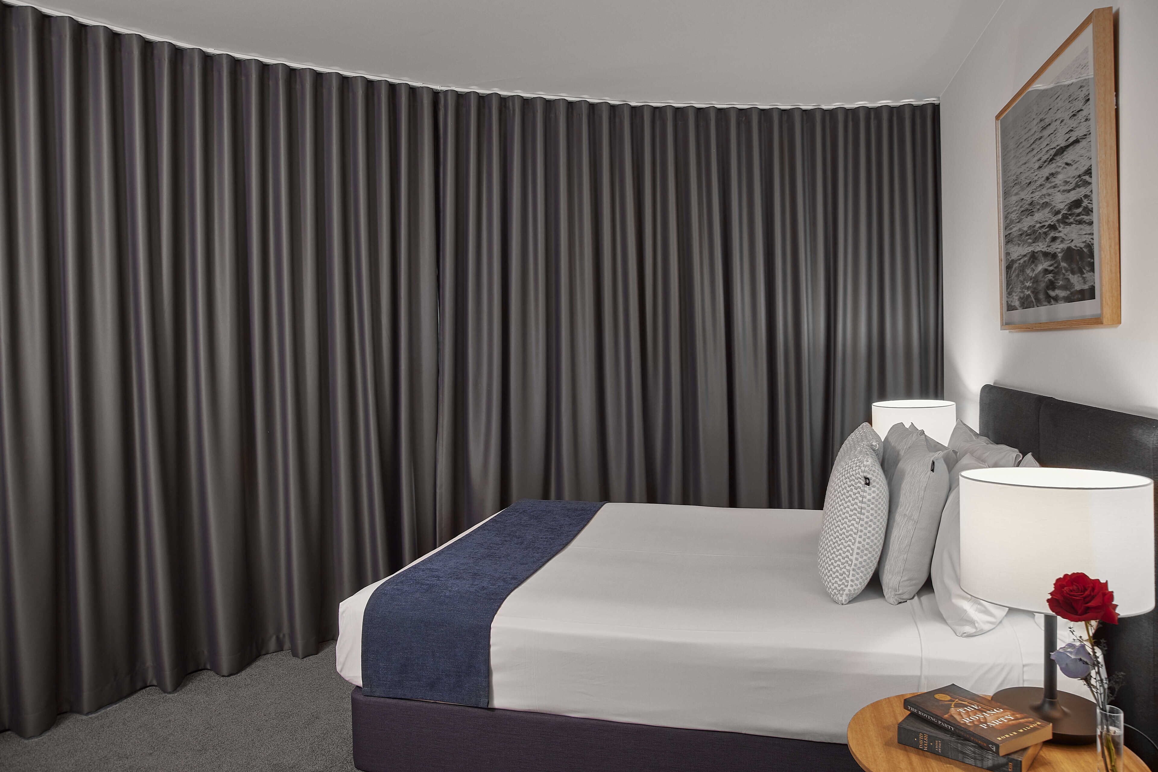 A hotel room with a large bed, grey curtains, a bedside lamp, a vase with a red rose, and a framed picture on the wall.