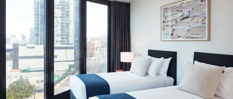A hotel room with two beds, a large window with a city view, a lamp, and a framed abstract painting.