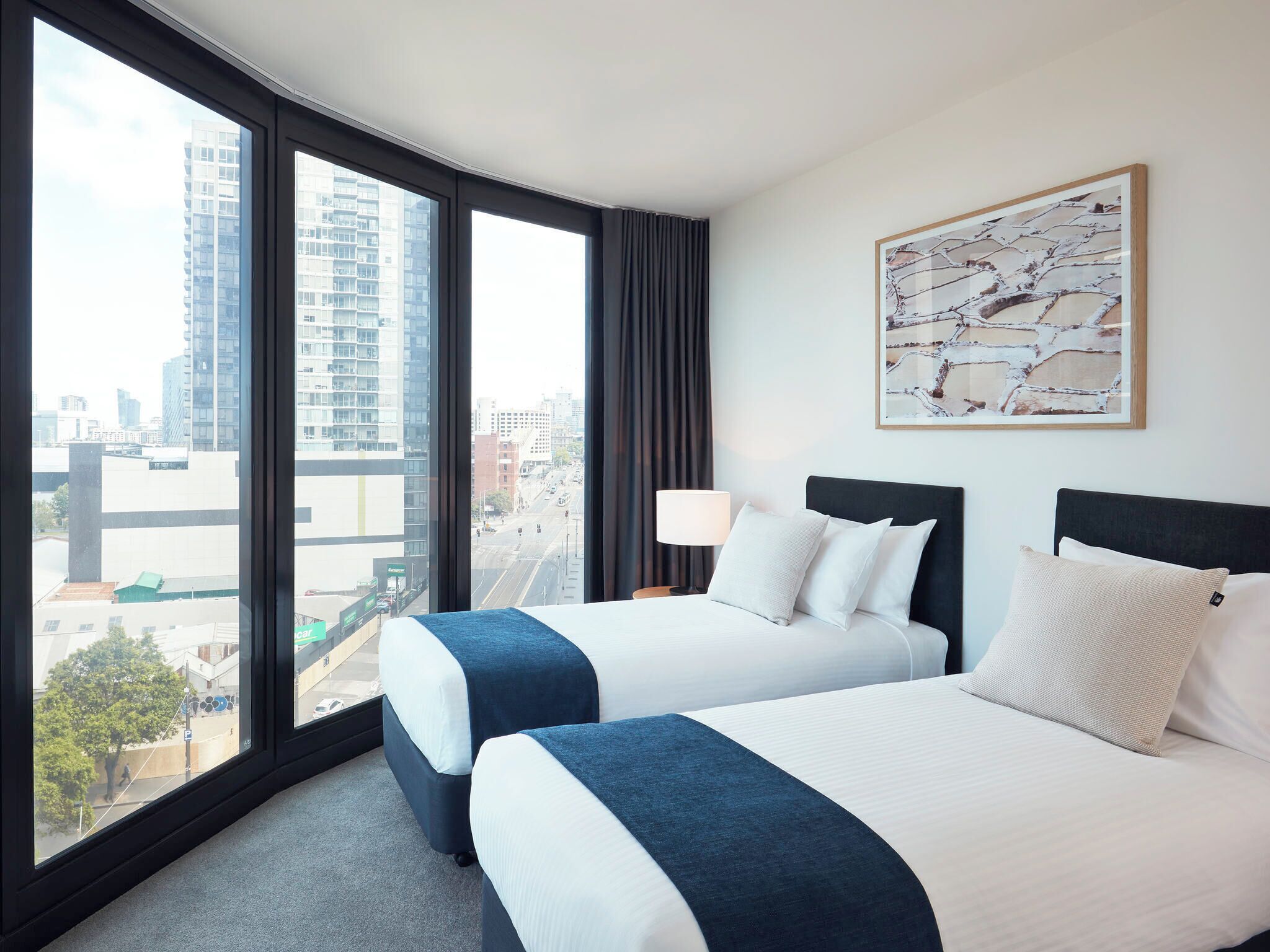 A hotel room with two beds, a large window with a city view, a lamp, and a framed picture on the wall.