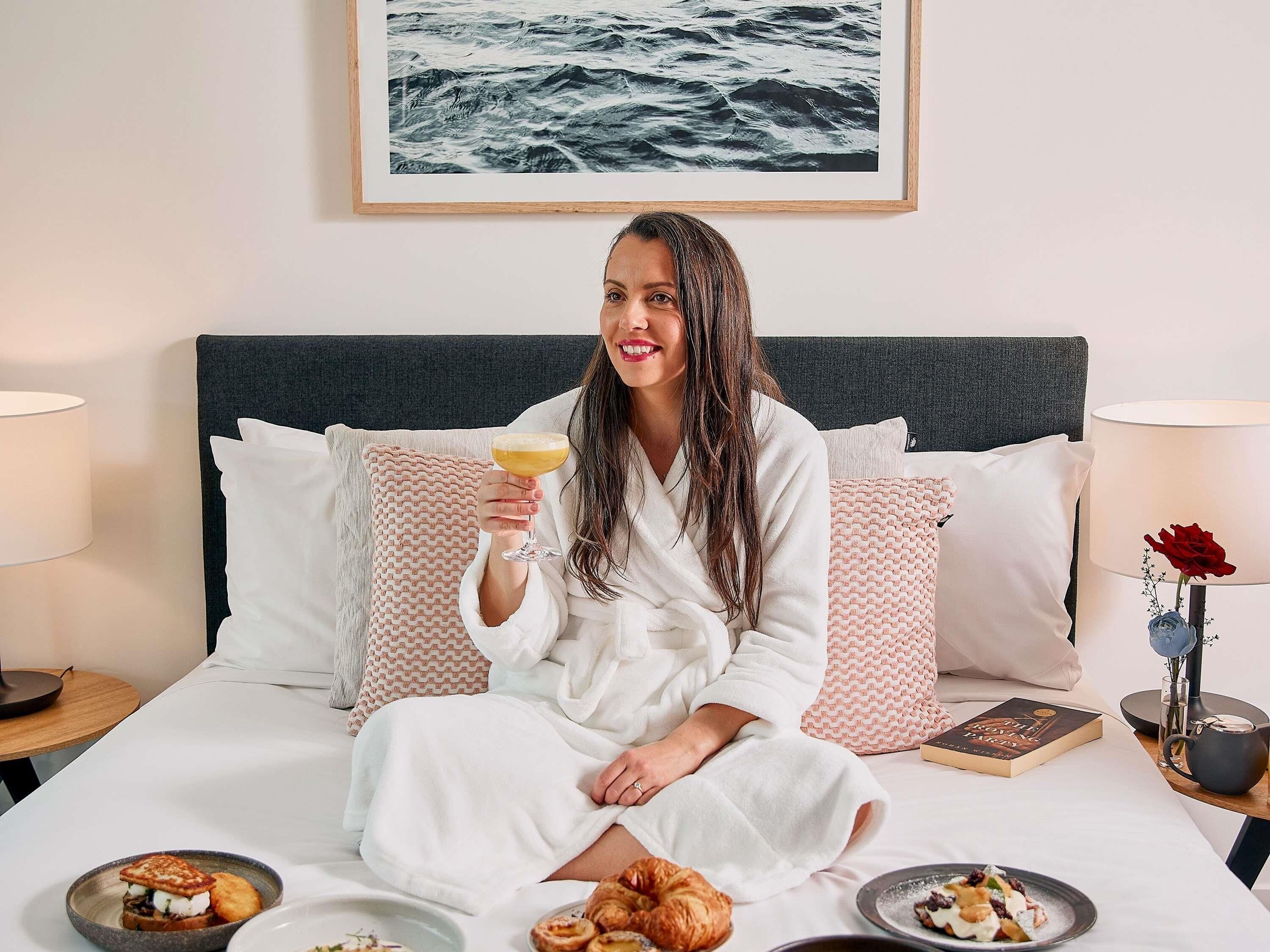 A person sitting on a bed with a drink, pillows, a book, and food items on the bedside table.