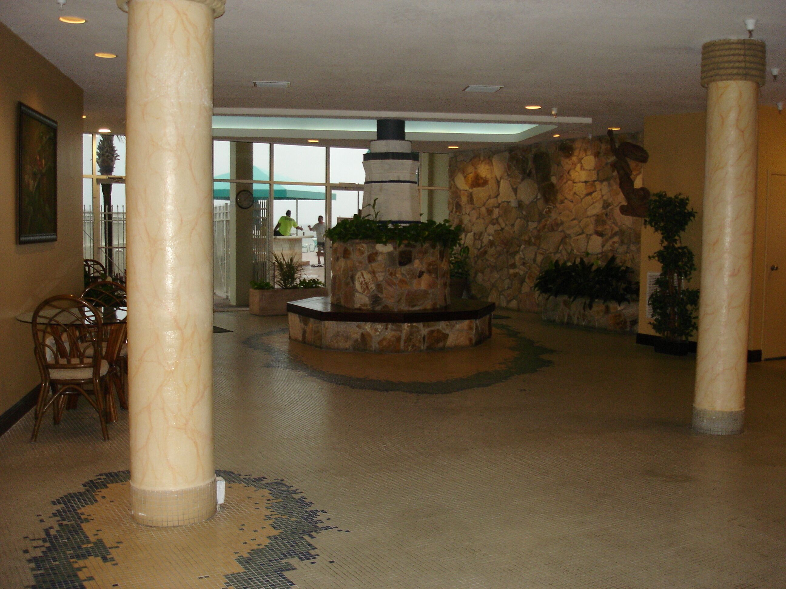 Een ruime lobby met een stenen muur, een centrale plantenbak met beplanting, en een zithoek met stoelen.
