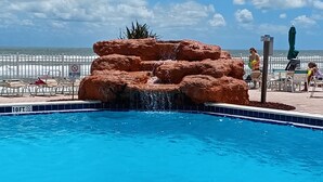 A pool with a rock waterfall feature, surrounded by lounge chairs and a white fence. - Gorgeous Ocean View & Heated Pool! Seasonal Discounts (Daytona Beach)