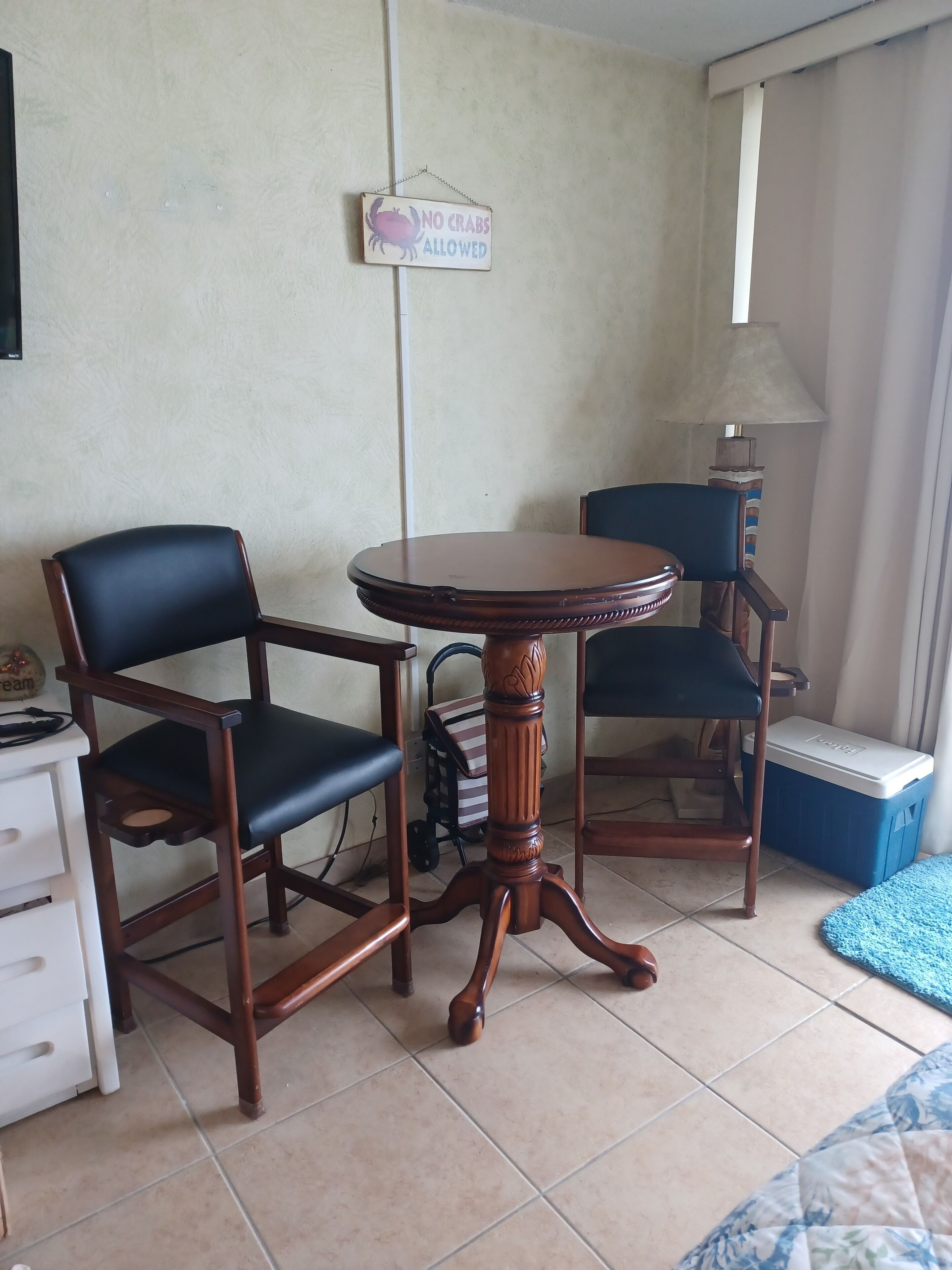 Une pièce comprenant deux chaises en bois, une petite table ronde, un bureau blanc, une lampe, une poubelle et un panneau accroché au mur.