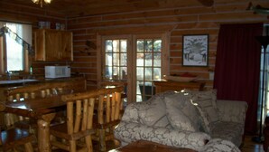 Interior de una cabaña de troncos con una mesa de comedor de madera, sillas, un sofá y una zona de cocina con microondas.