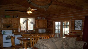 Interior de una cabaña con una mesa de comedor de madera, un sofá, una cocina con estufa y un ventilador de techo.