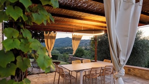 Um pátio externo com mesa e cadeiras de pedra, rodeado por vegetação e vista para o campo.