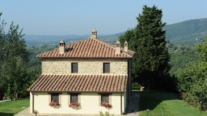 Uma casa de pedra de dois andares com telhado de cerâmica, rodeada por vegetação e colinas.