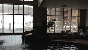 Une piscine intérieure agrémentée d’une cascade, de grandes fenêtres offrant une vue sur un espace extérieur enneigé, ainsi que plusieurs chaises et tables réparties dans la pièce.