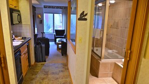 Un couloir étroit mène à une salle de bain équipée d’une baignoire, d’une douche et d’un lavabo. Une valise est posée sur le sol et une chaise se trouve au fond de la pièce.