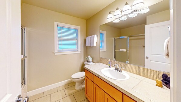 Une salle de bain équipée de toilettes, d’un lavabo, d’un miroir et d’une fenêtre avec des stores.