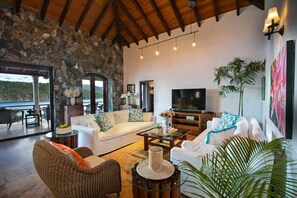A spacious living area with a stone wall, wooden ceiling, and a large flat-screen TV. There are multiple seating arrangements including a sofa, chairs, and a coffee table. - Blackbeard's Hideaway at Steele Point (West End)