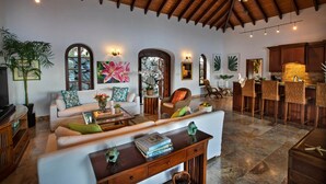 A spacious living area with a high ceiling, wooden beams, and a stone-tiled floor. There is a seating area with a sofa and armchairs, a coffee table, and a dining area with bar stools. - Blackbeard's Hideaway at Steele Point (West End)