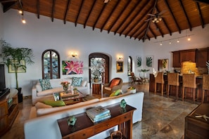 A spacious living area with a high ceiling, wooden beams, and a stone-tiled floor. There is a seating area with a sofa and armchairs, a coffee table, and a dining area with bar stools.
