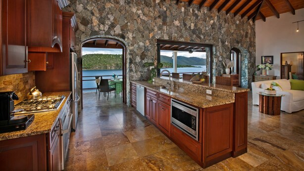 A kitchen with a stone wall, wooden cabinets, granite countertops, and a view of the ocean. - Blackbeard's Hideaway at Steele Point (West End)