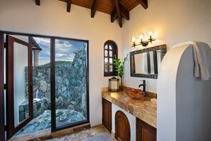 A bathroom with a stone wall, a wooden door, a sink with a copper basin, and a mirror with lights. - Blackbeard's Hideaway at Steele Point (West End)