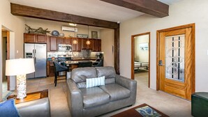 A modern kitchen with dark wooden cabinets, a stainless steel refrigerator, and a microwave. There is a dining area with a black chair and a table. A gray leather sofa is in the living area with a lamp and a rug. - Shadow Palm Villa 2 Private Resort Villa (Port Aransas)