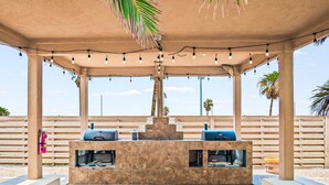 An outdoor patio with a stone barbecue area, string lights, and a view of palm trees. - Shadow Palm Villa 2 Private Resort Villa (Port Aransas)