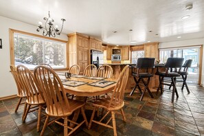 A dining area with a wooden table and chairs, a chandelier, and a kitchen in the background. - Bear mountain resort 5bd 3 bath sleeps 2600 sq (Big Bear Lake)
