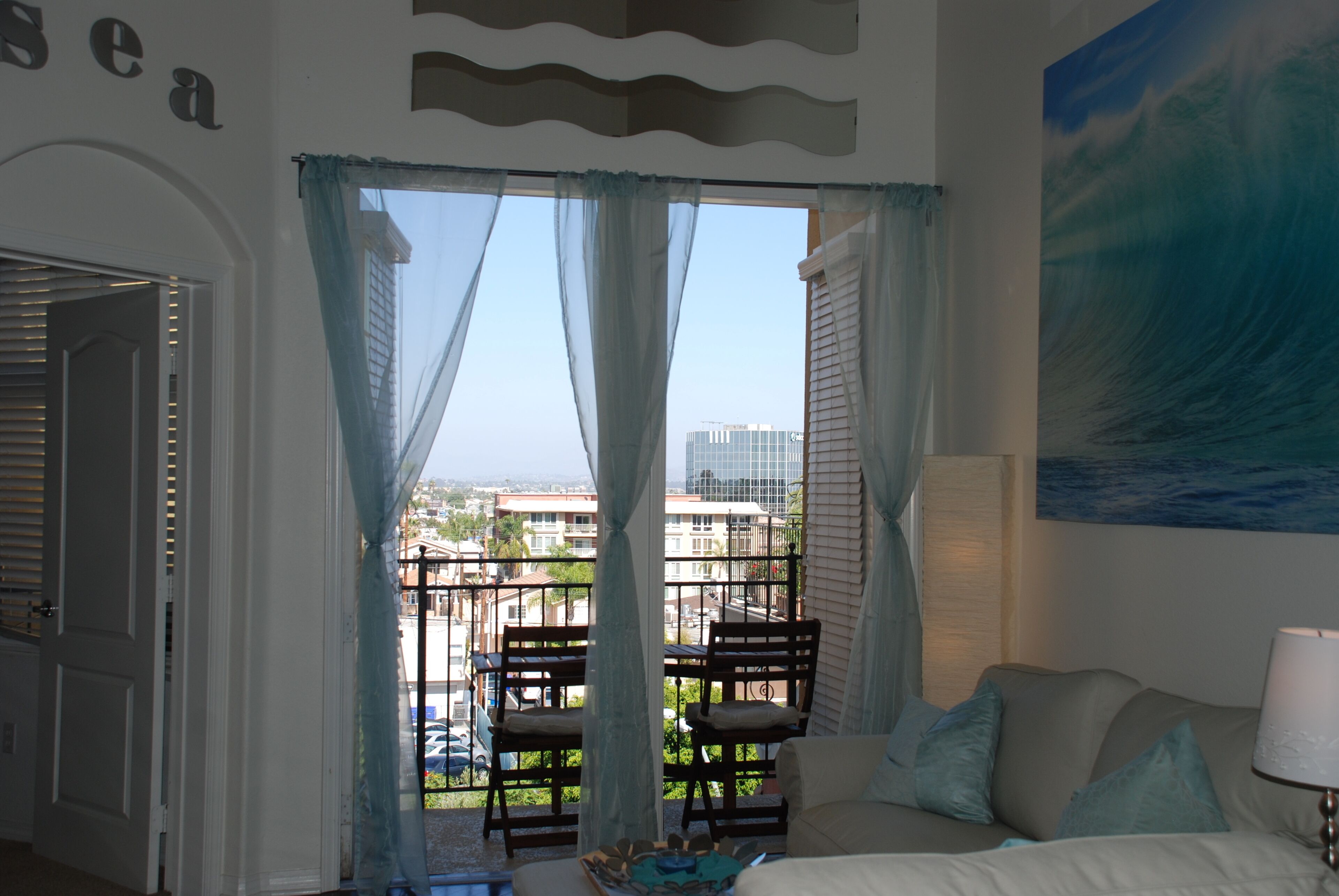 A room with a balcony, a bed, a chair, and a painting of a wave.