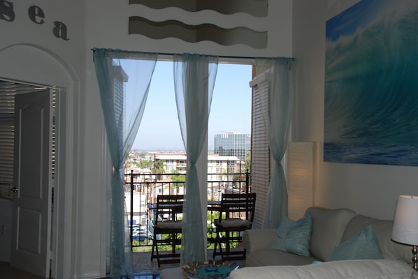 A room with a balcony, a bed, a chair, and a painting of a wave.