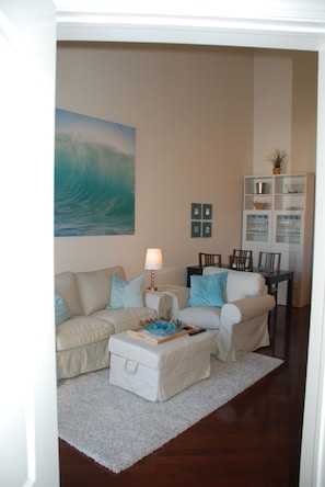 A living room with a beige sofa, a white ottoman, a coffee table, a lamp, a painting of a wave, and a dining area with chairs and a cabinet.