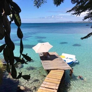 A wooden dock leading to a beach with clear water, a boat, and an umbrella. - walk to the Beach, Playa Santa  or playa monjas Apt with paddleboard and kayak (Ensenada)