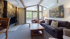 A cozy living room with a stone fireplace, a wooden coffee table, a sofa, and a large window offering a view of the outdoors.