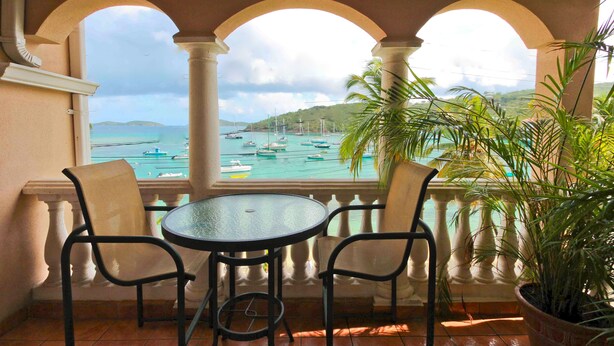 A balcony with two chairs and a table, overlooking a marina with boats and a hilly landscape. - Grande Bay SURFSIDE Studio, Beachfront, Perfect View/Location (Cruz Bay)
