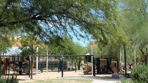 Children can play on swings, slides, and climbing elements in a shaded playground area surrounded by trees.