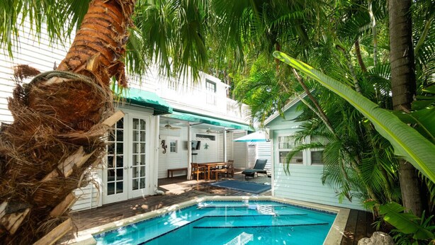 Un área de piscina con cubierta de madera, un espacio de estar techado y un edificio blanco con puertas de cristal.