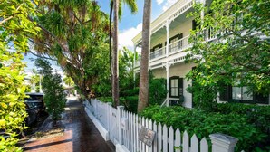Un estrecho sendero de ladrillo bordeado por una cerca blanca de piquetes y abundante vegetación, que conduce a un edificio de estilo colonial blanco con balcones.