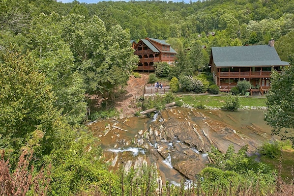A lodge with a wooden exterior, a bridge over a stream, and a forested backdrop.