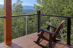A wooden deck with a metal railing, a bench, and a view of a forested area with mountains in the distance.