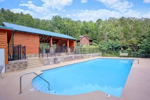 A swimming pool with a stone wall and metal railings, surrounded by a wooden cabin and a second building with a metal roof.