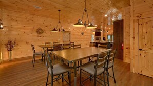 A wooden interior with a large dining table surrounded by chairs, a kitchen area, and a wooden door.