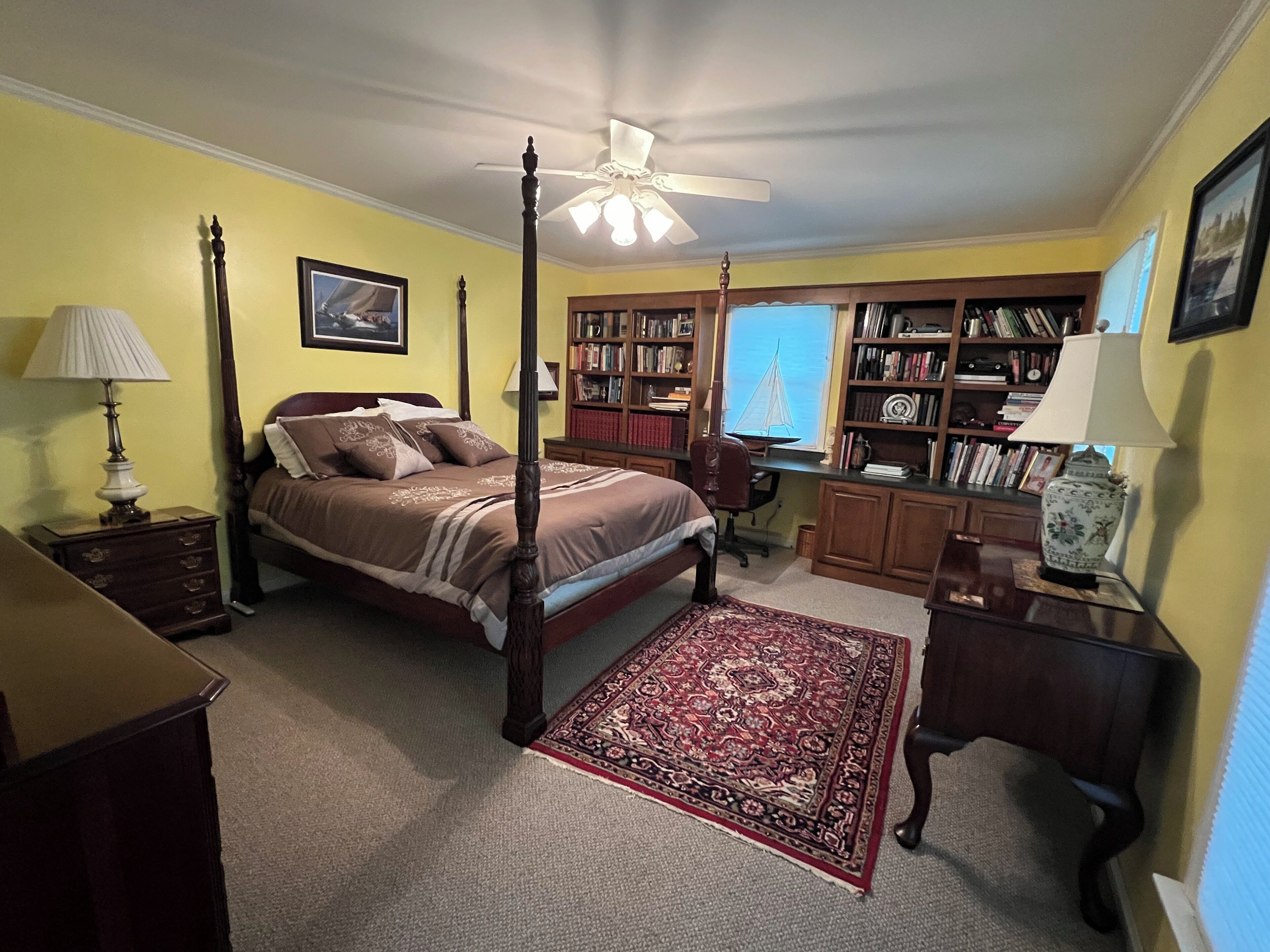 A bedroom with a four-poster bed, a ceiling fan, a bookshelf, and a desk with a chair.