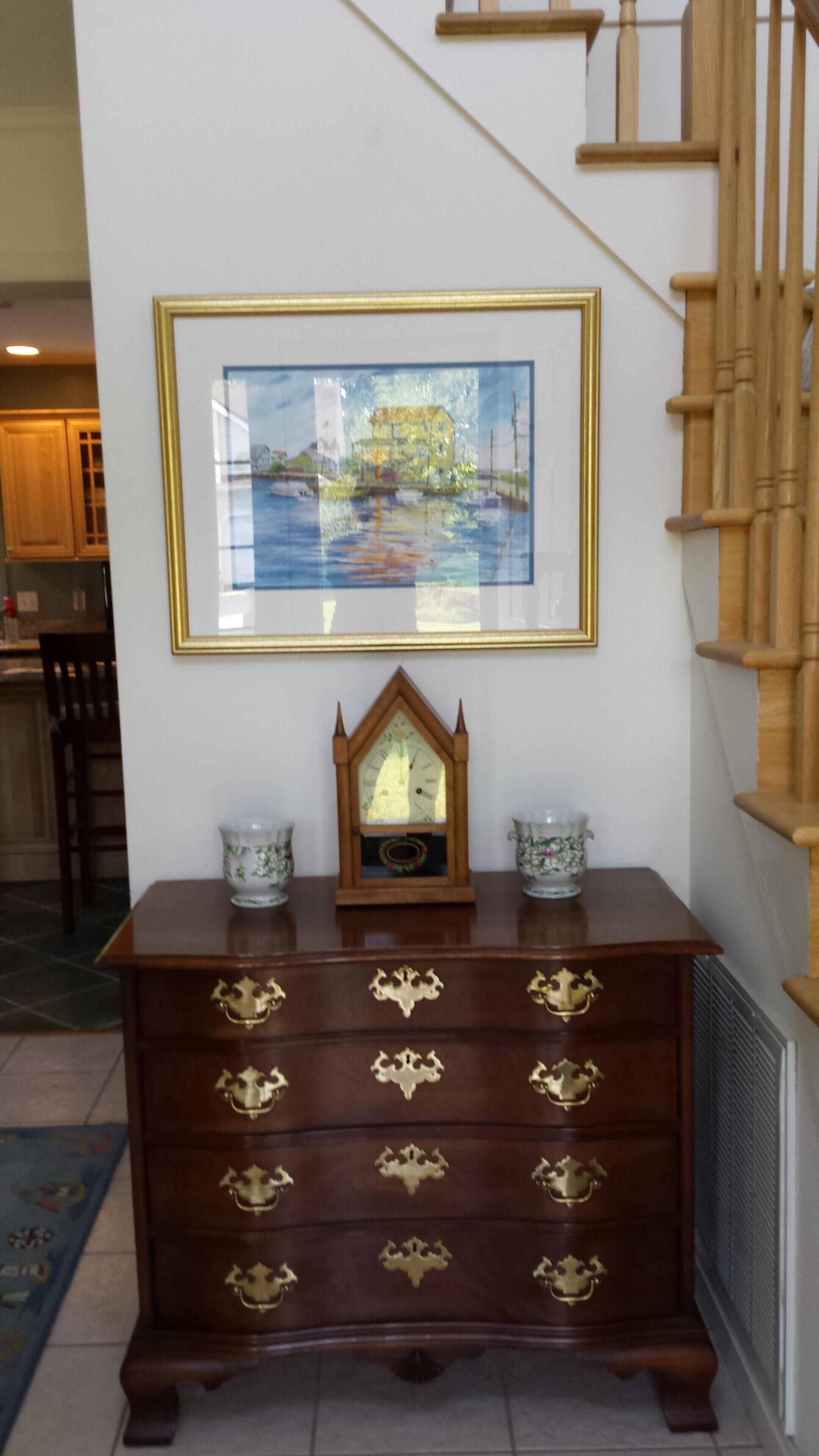 A wooden dresser with brass handles, a framed painting above it, and a wooden staircase to the right.