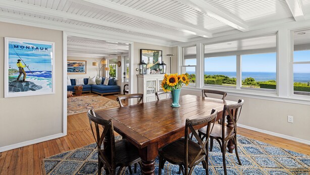 Un coin repas avec une table et des chaises en bois, un vase rempli de tournesols et une vue sur l’océan par de grandes fenêtres.